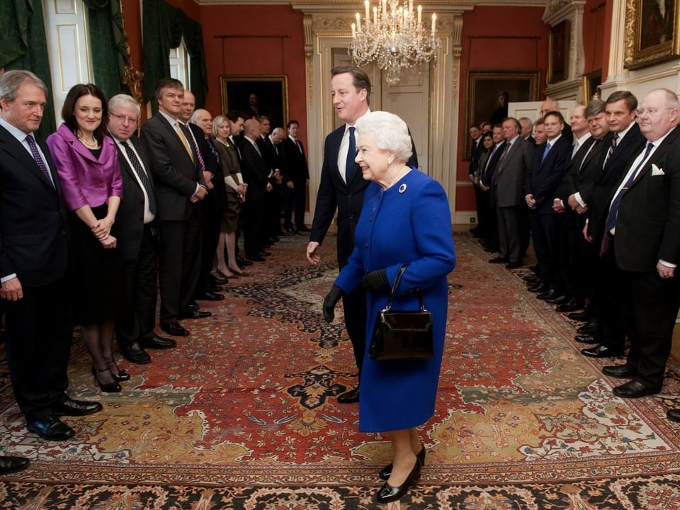 Die Queen im blauen Kleid steht mit einem Mann in der Mitte des Raumes. Der Boden ist mit einem wertvoll aussehenden Teppich belegt. An den Wänden haben sich Leute in Anzügen in Reih und Glied aufgestellt.