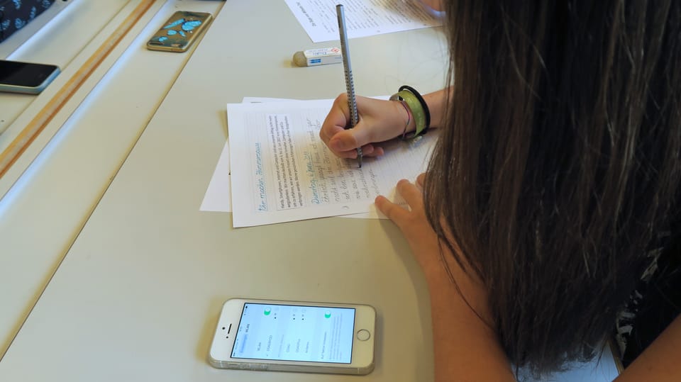 Ein Mädchen mit langen Haaren schreibt Schularbeiten auf dem Tisch liegt ein Handy. 