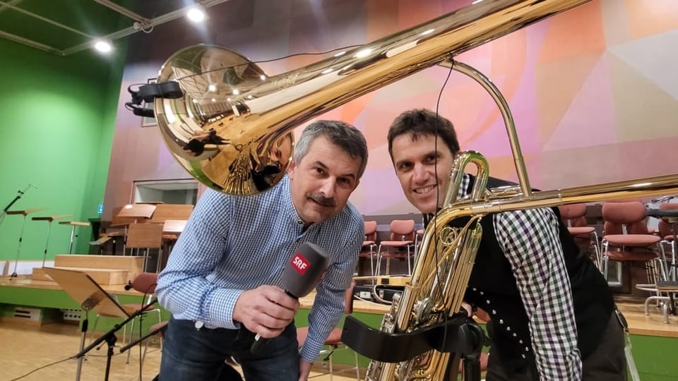 Zwei Männer mit Mikrofon in der Hand halten ein riesiges Blasmusikintstrument. 