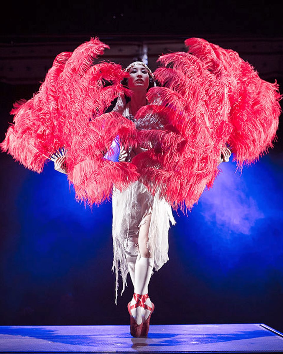 Eine Frau steht auf der Spitze von roten Ballettschuhen und hält zwei riesige rote Federfächer vor sich.