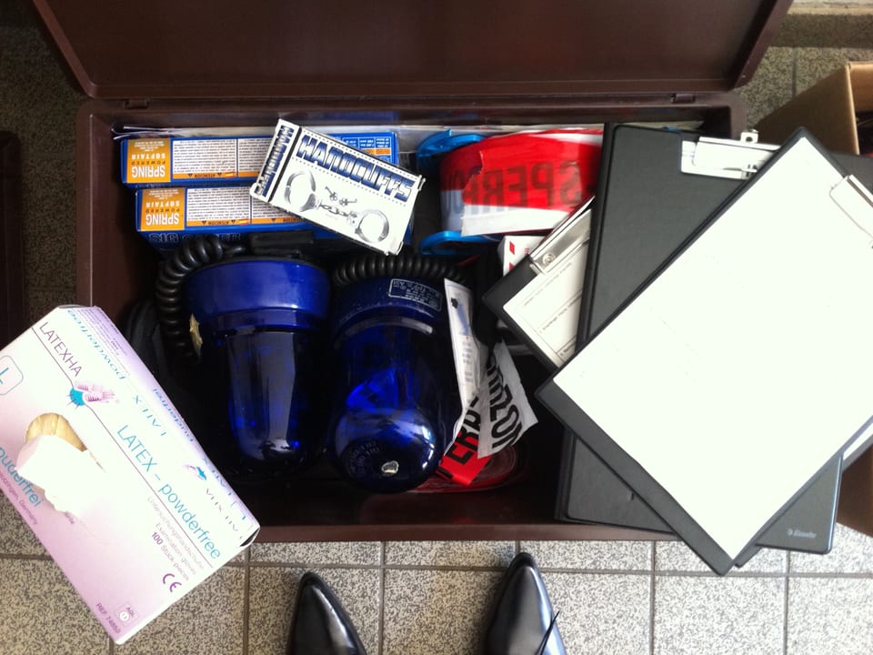 Ein Koffer gefüllt mit Polizei-Utensilien: Ein Blaulicht, Absperrband, Handschellen und Latex-Handschuhe.