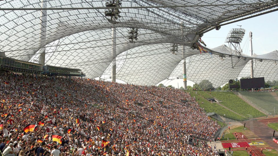 Ein zeltartiges Dach des Olympiastadions in München, Fans auf der Tribüne.