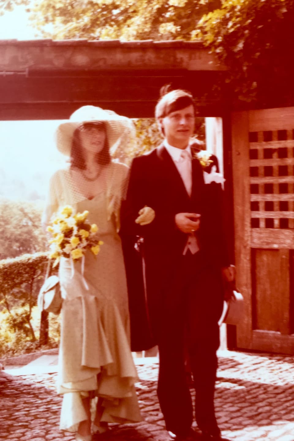 Das Brautpaar, er im Frack, ssie im scheinbar crèmefarbenen Kleid und mit gelbem Blumenstrauss steht Arm in Arm da.