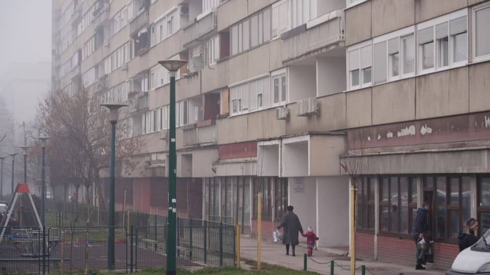 Eine Frau geht mit einem Kind an der Hand die Strasse vor einem Block entlang.