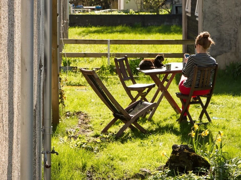 Eine Frau sitzt im Garten und schaut auf ihr Smartphone. Auf dem Tisch sitzt eine Katze.