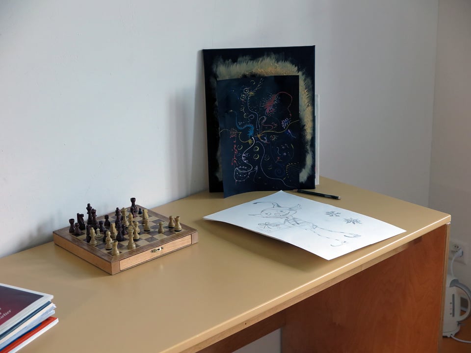 Auf einem Pult stehen ein kleines Schachbrett, eine Zeichnung mit einer Fantasiefigur und ein farbiges Bild.