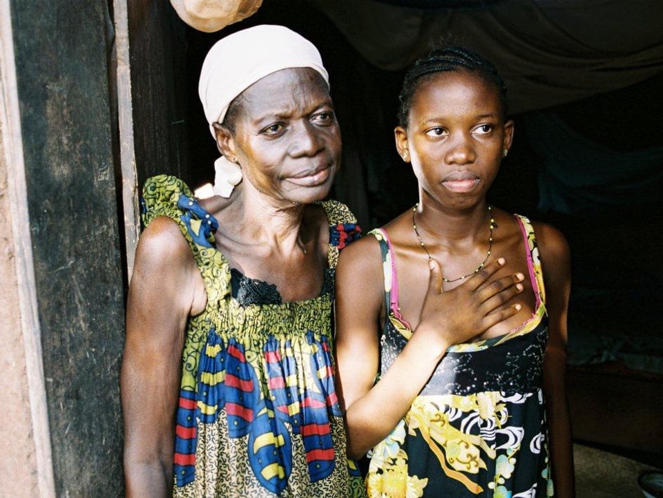Eine ältere Afrikanerin und ein junges Mädchen stehen in der Tür eines Hauses. Beide schauen an der Kamera vorbei aus de, Bild heraus, das Mädchen hat die rechte Hand auf die Brust gelegt.