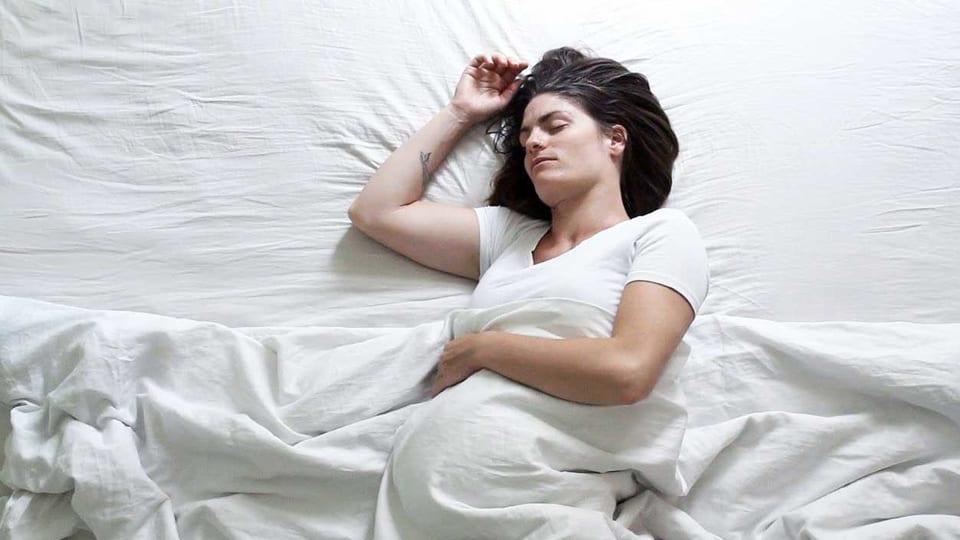 Eine schwangere Frau liegt im Bett. Das Laken und die Matraze sind weiss.