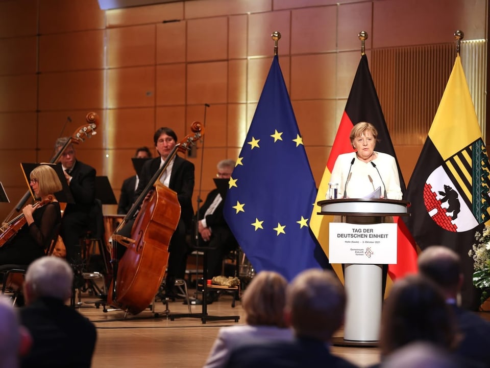 Angela Merkel an einem Rednerpult. Im Hintergrund Flaggen und Musiker.