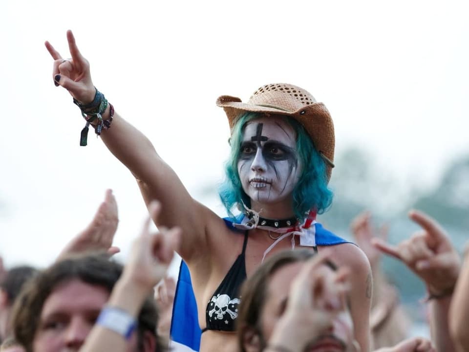 Eine Schwarz Weiss geschminkte Frau im Publikum eines Metal-Konzerts streckt die gehörnte Hand in die Luft.
