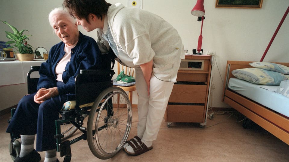 Eine Pflegerin beugt sich vor zu einer älteren Dame die im Rollstuhl sitzt und etwas erzählt, die beiden befinden sich ein einem Zimmer im Altersheim.