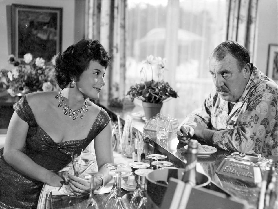 Ein beleibter Mann mit Hawaii-Hemd lehnt an einer Getränke-Theke. Dahinter steht eine elegant gekleidete Frau, die ihn anlächelt und ein Glas in der Hand hält.