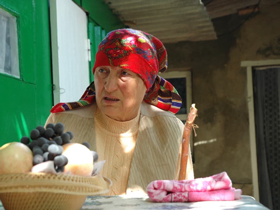 Eine alte Frau mit Kopftuch sitzt an einem Tisch.