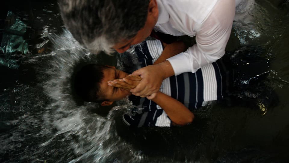 Ein Mann taucht ein Kind ins Wasser. Es handelt sich um eine Taufe.