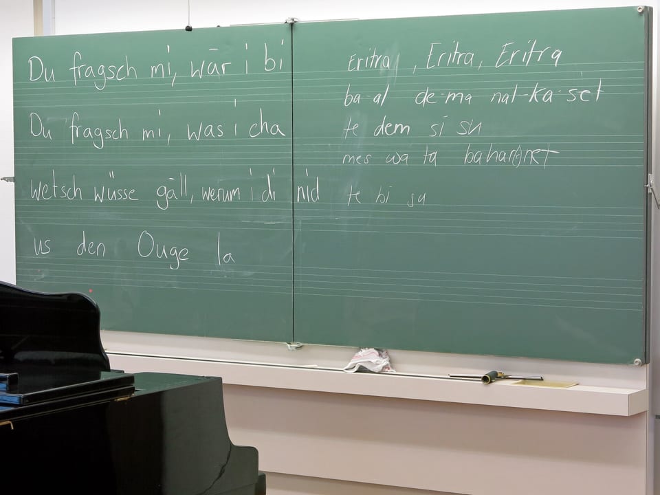 Eine grüne Wandtafel mit berndeutschen und eritreischen Liedtexten hinter einem Klavier.