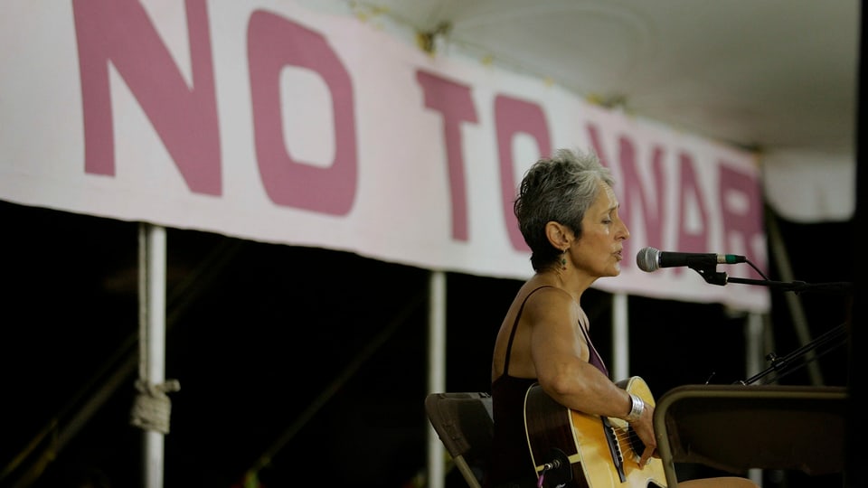 Frau mit kurzen gräulichen Haaren sitzt mit Gitarre auf einer kleinen Bühne und singt in ein Mikrofon