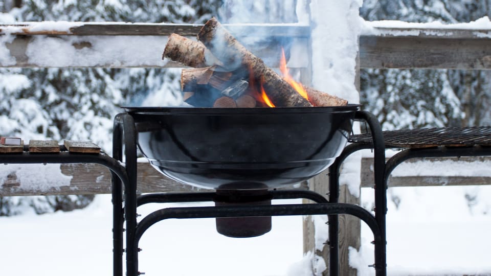 Ein Grill mit brennenden Holzscheiten steht in einem Wald im Schnee.