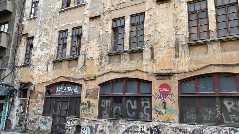Ein vergilbter Wohnblock: der Verputz bröckelt überall ab, unten voller Graffiti, und ein roter Punkt... 