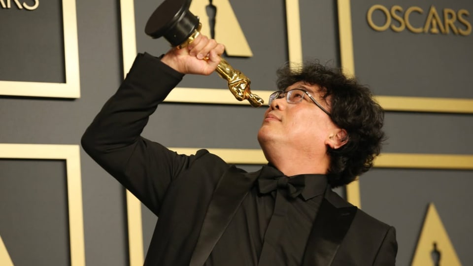 koreanischer Mann mit wuscheligen Haaren und Brille in schwarzem Anzug, hält Oscar in angewinkeltem Arm.
