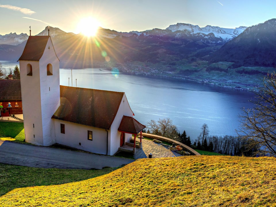 Kapelle hoch über dem See und dahinter Sonnenaufgang über den Alpen