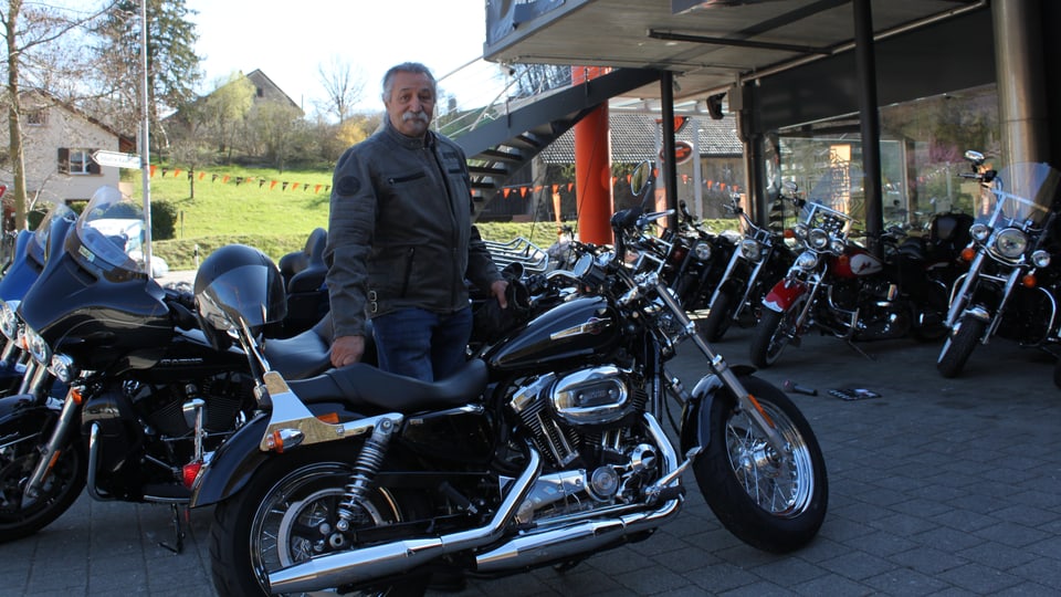 Mann steht hinter seiner schönen Harley - Im Hintergrund Töffladen zu erkennen