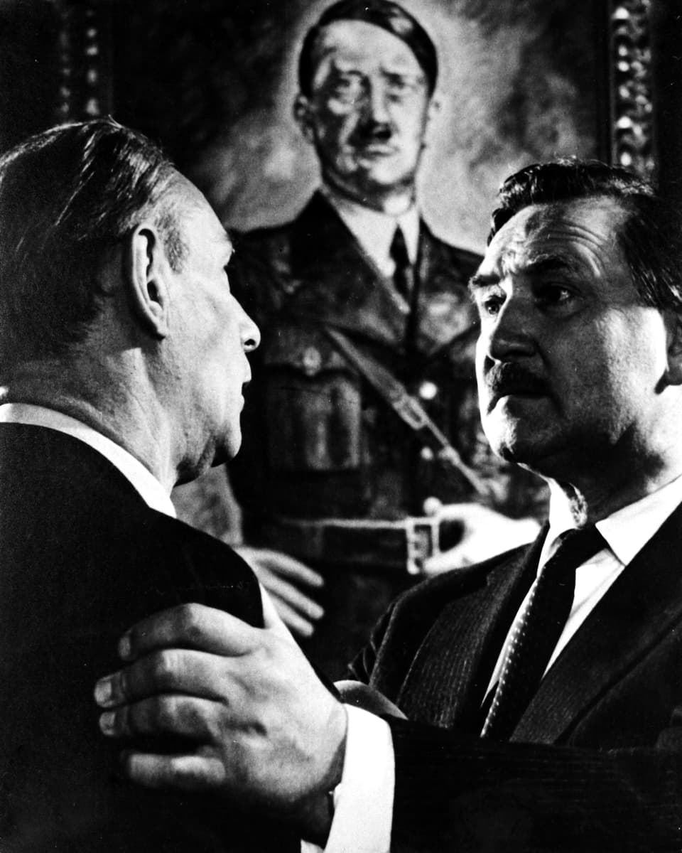 Zwei Männer stehen sich gegenüber. Der eine hält den anderen bei den Schultern. Im Hintergrund hängt ein Bild Hitlers.