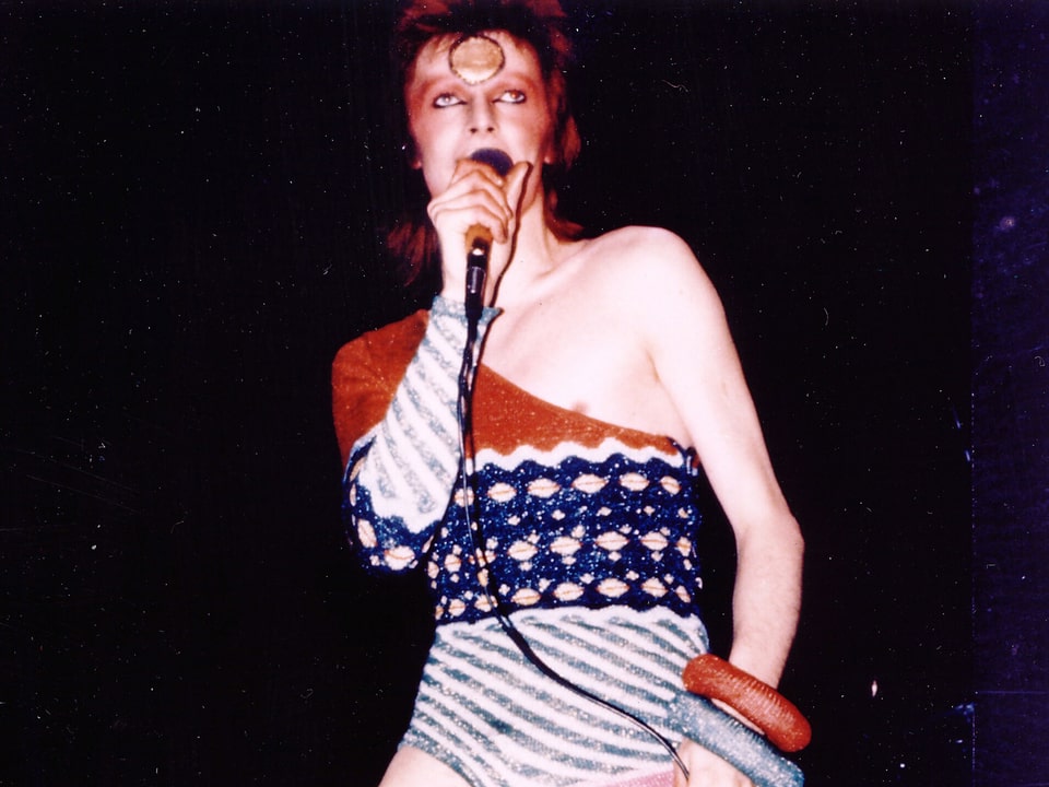 David Bowie in engem, farbigen Anzug, geschminkt.