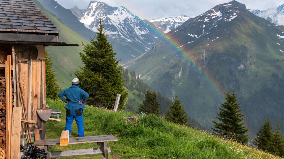 Eine Frau steht vor einer Alphütte und betrachtet einen Regenbogen.