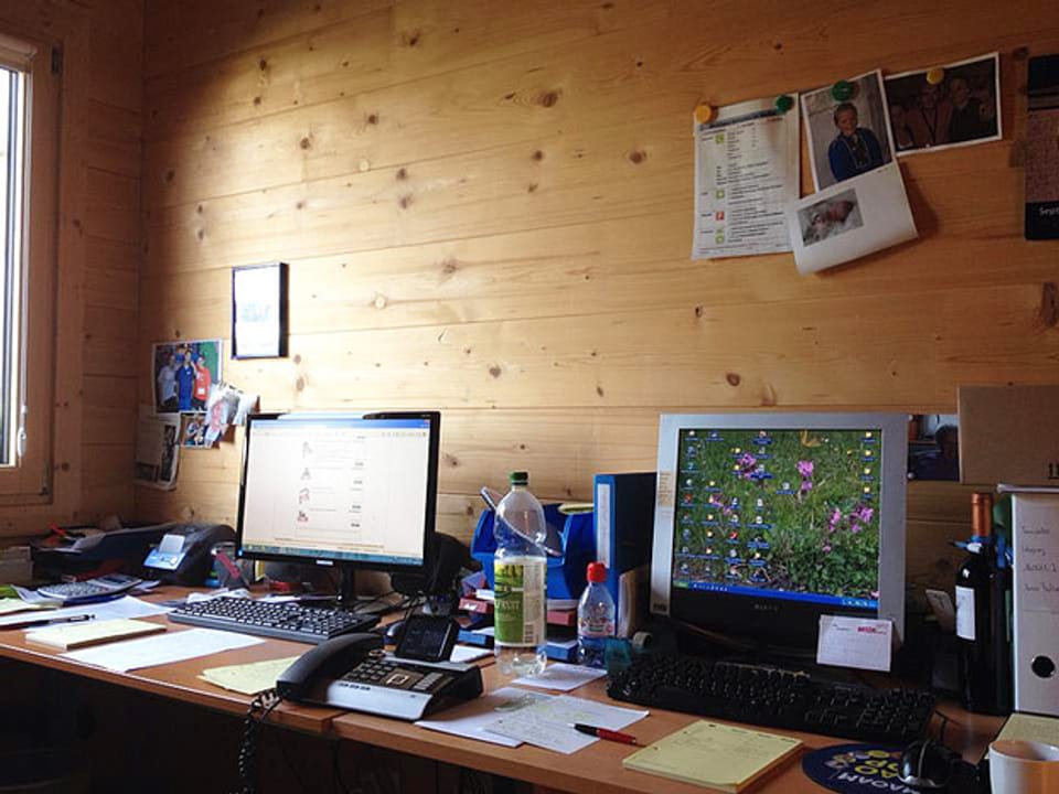Ein Schreibtisch mit zwei Computern und vielen losen Blättern, einem Telefon und PET-Flaschen. Fotos an der Holzwand.