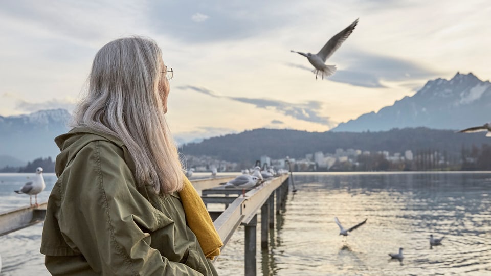 Eine Frau mit langen, grauen Haaren steht am Ufer eines Sees und schaut in die Ferne.