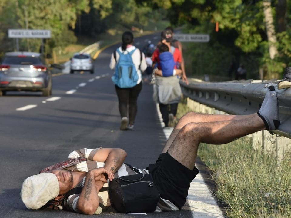 A migrant lies in the street and sleeps.  His face is covered by a hat.
