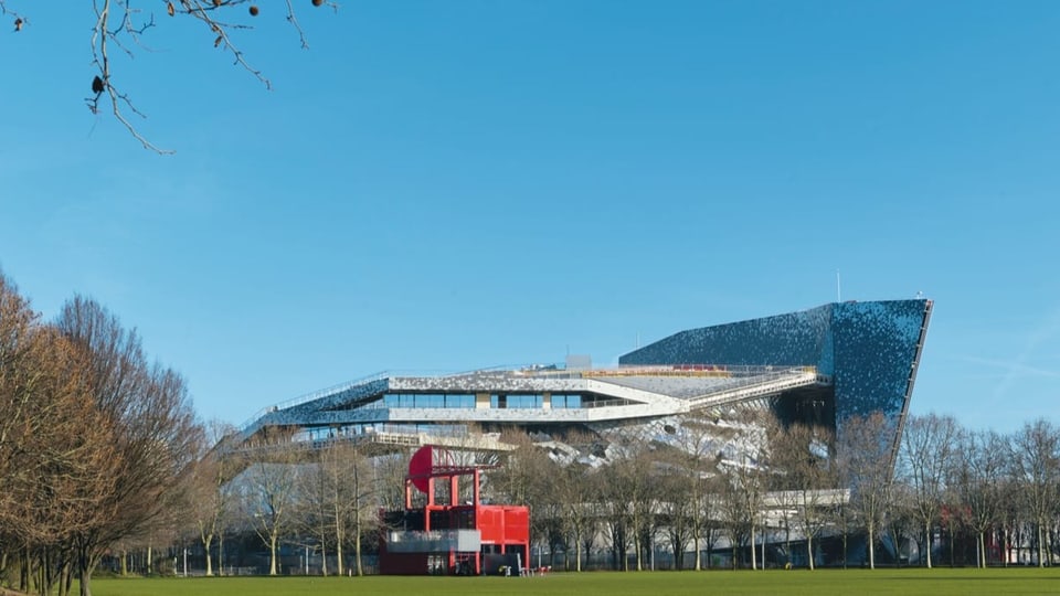 Ein grosses, silbernes Gebäude bei blauem Himmel, grüne Wiese und Bäume im Vordergrund.