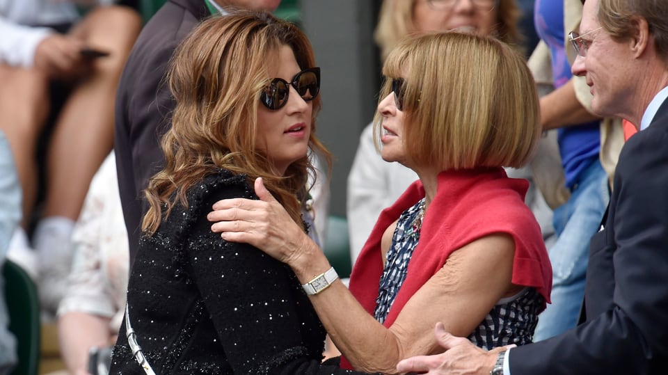 Mirka Federer und Anna Wintour geben sich begrüssung Küsschen. Federer trägt ein schwarzes Oberteil und eine dunkle Sonnenbrille, Wintour hat sich einen roten Pullover um die Schultern geschlungen.