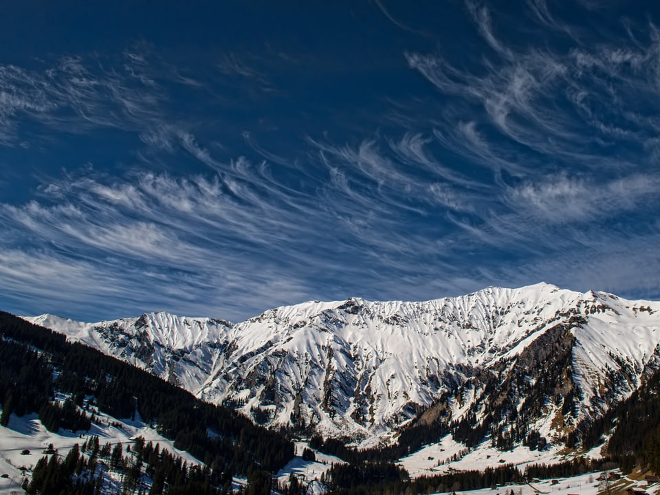 Blauer Himmel oben, unten verschneite Bergwelt. Am Himmel sind viele haarängliche, feine Wolken zu sehen. Sie haben hackenförmige Strukturen. 
