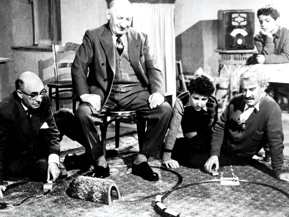 Männer und Jungen sitzen in einem Zimmer um eine Spielzeugeisenbahn herum und verfolgen amüsiert die Bahnfahrt der Spielzeuglokomotive.