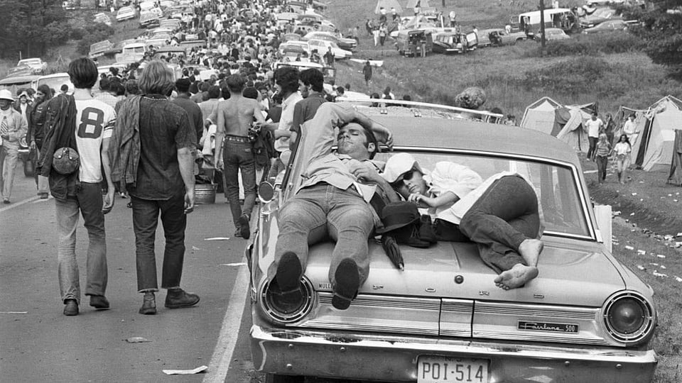 Ein Paar schläft auf der Kühlerhaube, im Hintergrund eine mit Autos und Menschen verstopfte Strasse
