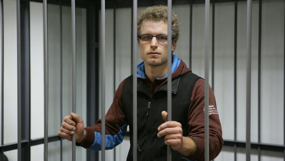 Marco Weber ist in einem Käfig eingesperrt. Er hat die Hände an den Gitterstäben und blickt in die Kamera.
