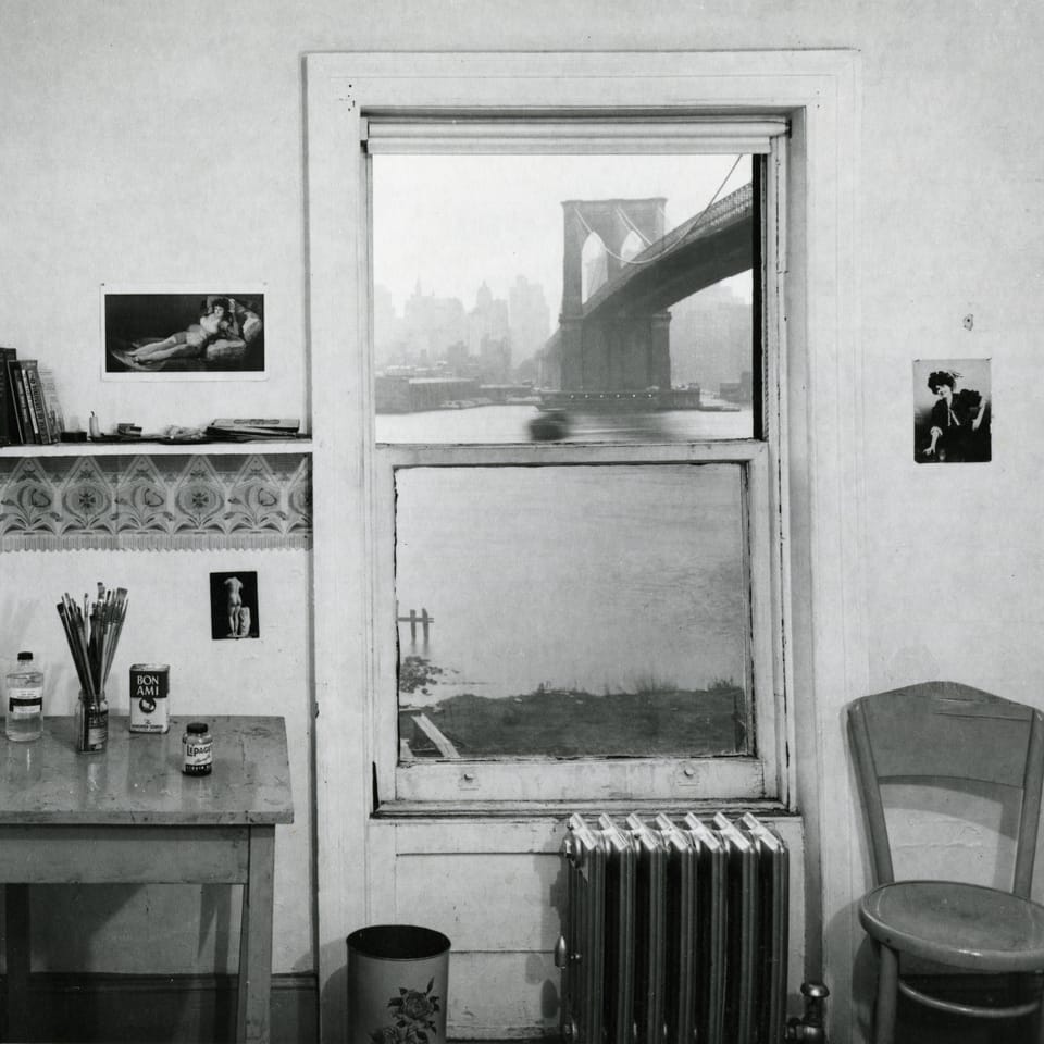 Auf einer schwarz-weissen Fotografie ist ein karg eingerichtetes Zimmer zu sehen, durch dessen Fenster die Brooklyn Bridge zu sehen ist. 