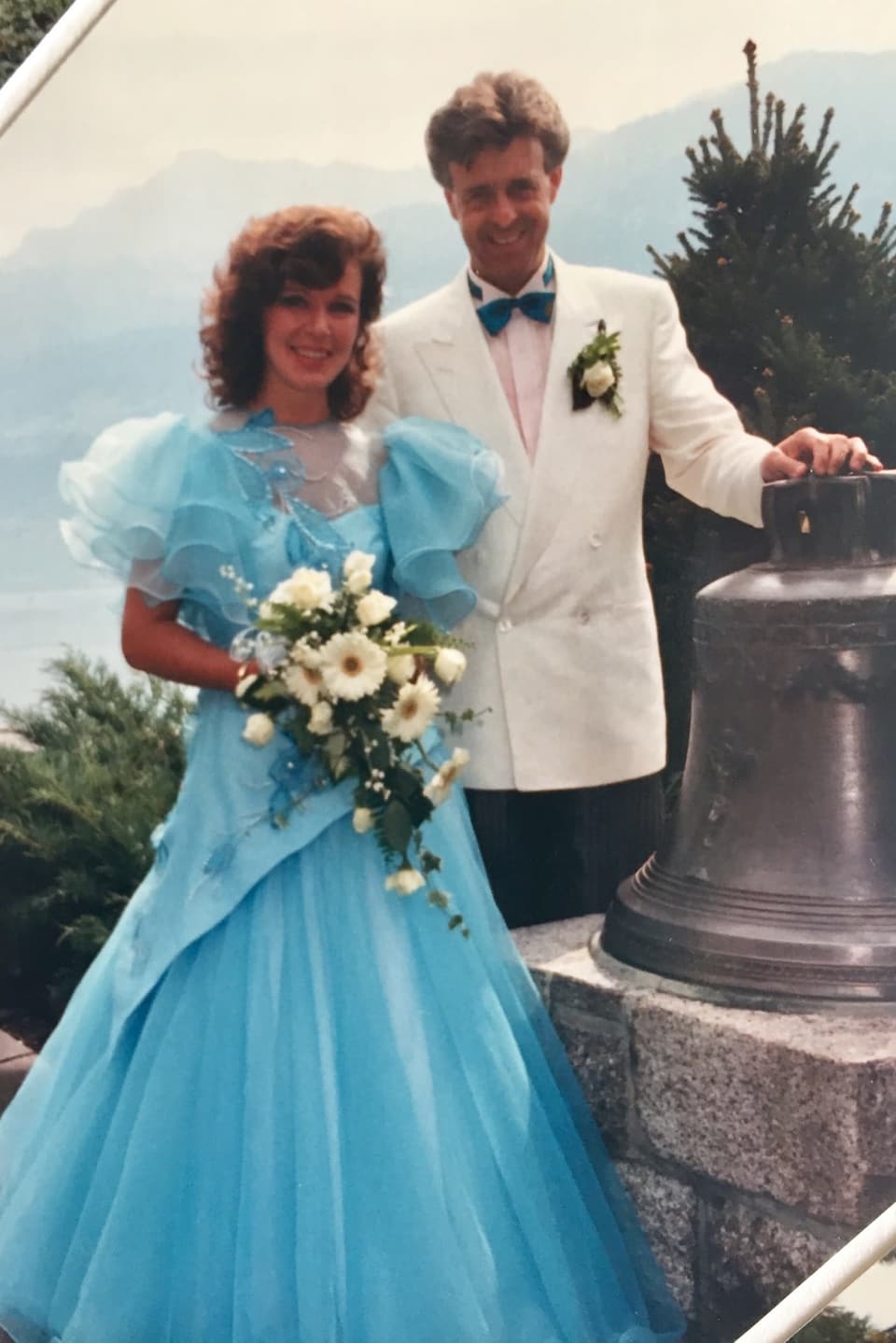 Ein Brautpaar, sie in einem hellblauen Kleid und er im weissen Blazer, stehen bei einer Glocke und lächeln in die Kamera. Im Hintergrund ein See.