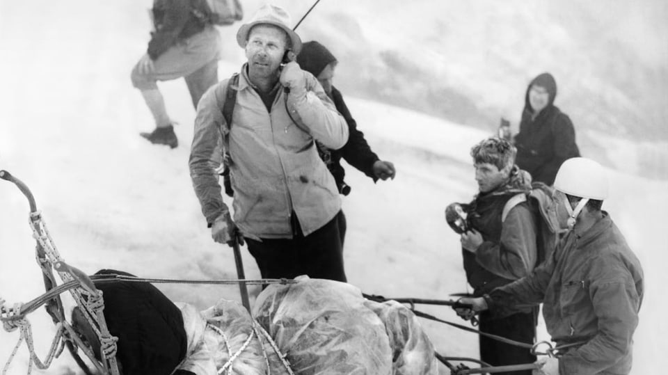 Schwarzweissfoto: Männer in Bergstigerkleidung neben einem Rettungsschlitten. Eer der Männer hält ein Telefon und blickt nach oben.