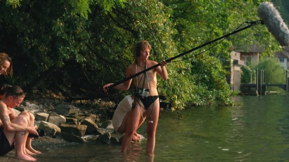Eine Frau mit entblösstem Oberkörper steht im Wasser eines Flusses und hält ein grosses Mikrofon an einer Stange.