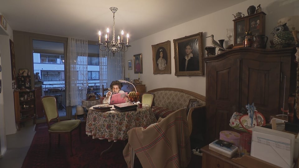 Frau sitzt in einem Wohnzimmer, das mit alten Möbeln und Bildern vollgestellt ist.
