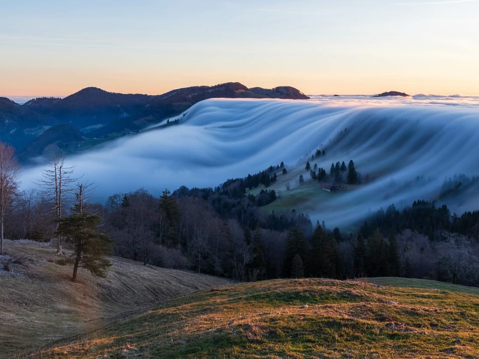 Hügelige Landschaft mit Nebel rechts, der wie ein Wasserfall nach linkls strömt und sich auflöst. 