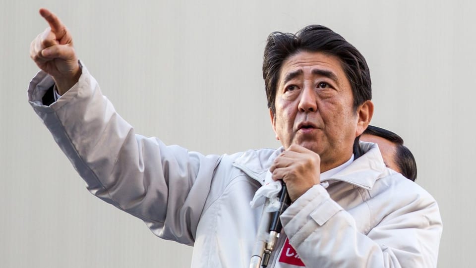 Shinzo Abe hält ein Mikrofon und hält den rechten Arm mit ausgestrecktem Zeigefinger hoch