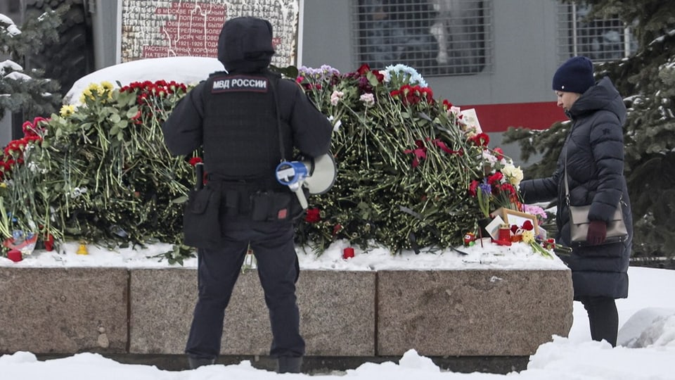 Eine Frau legt Blumen nieder, neben ihr steht ein Polizist. 