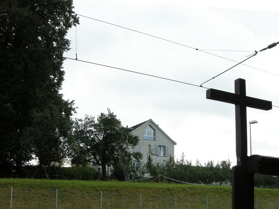 Blick aus der Ferne auf ein alleinstehendes weisses Wohnhaus, im Vordergrund ist ein grosses Holzkreuz zu sehen.