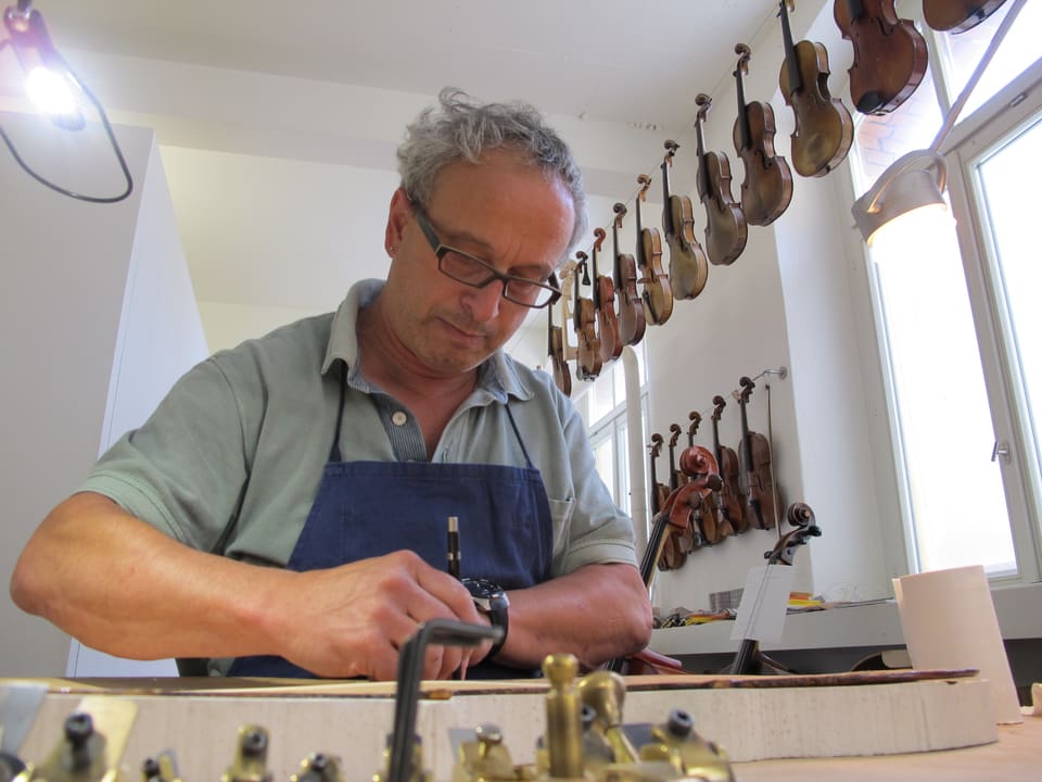 Der Geigenbauer arbeitet an einem Cello, im Hintergrund hängen mehrere Geigen.