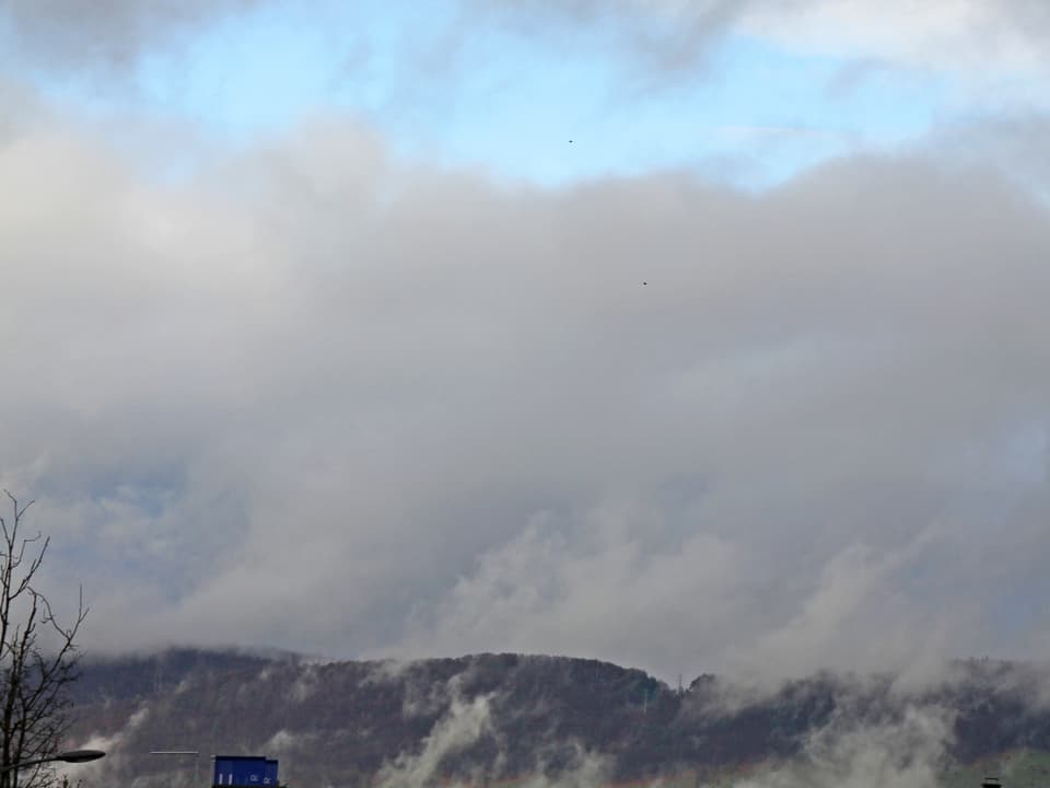 Unten sieht man einen bewaldeten Hügelzug. Darüber liegt eine dicke, graue Wolke. Allerdings zeigt sich am oberen Bildrand eine blaue Aufhellung.