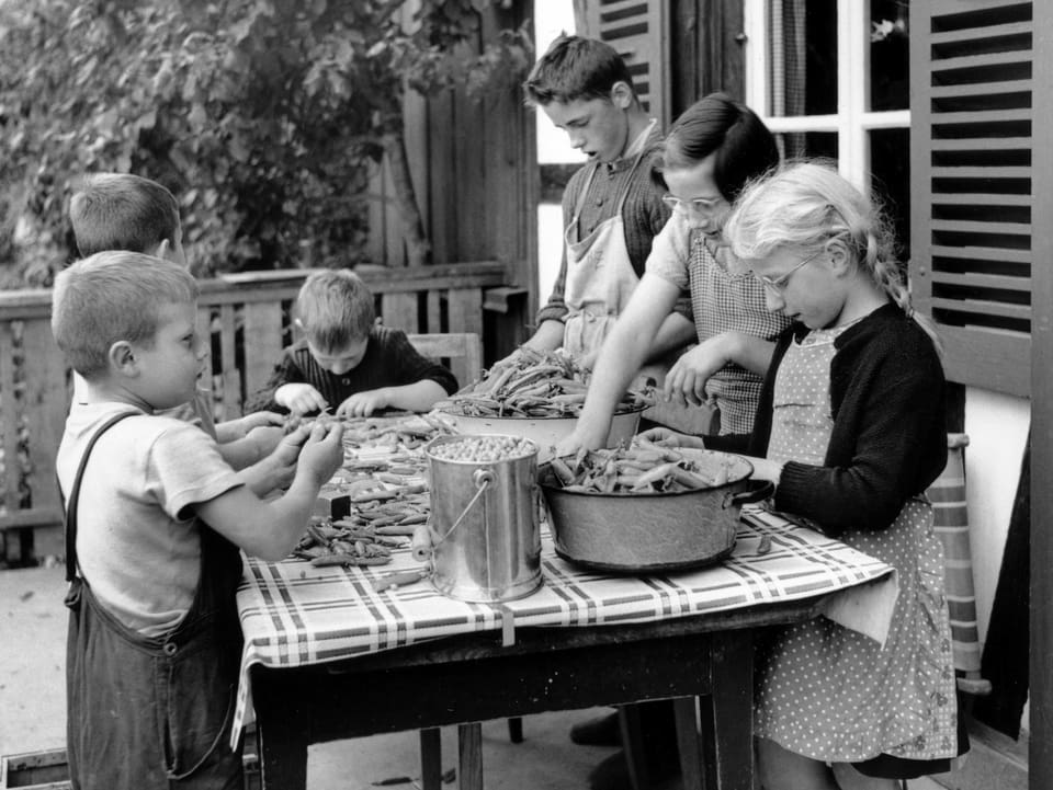 Kinder sitzen draussen um einen Tisch und bereiten Bohnen zu.