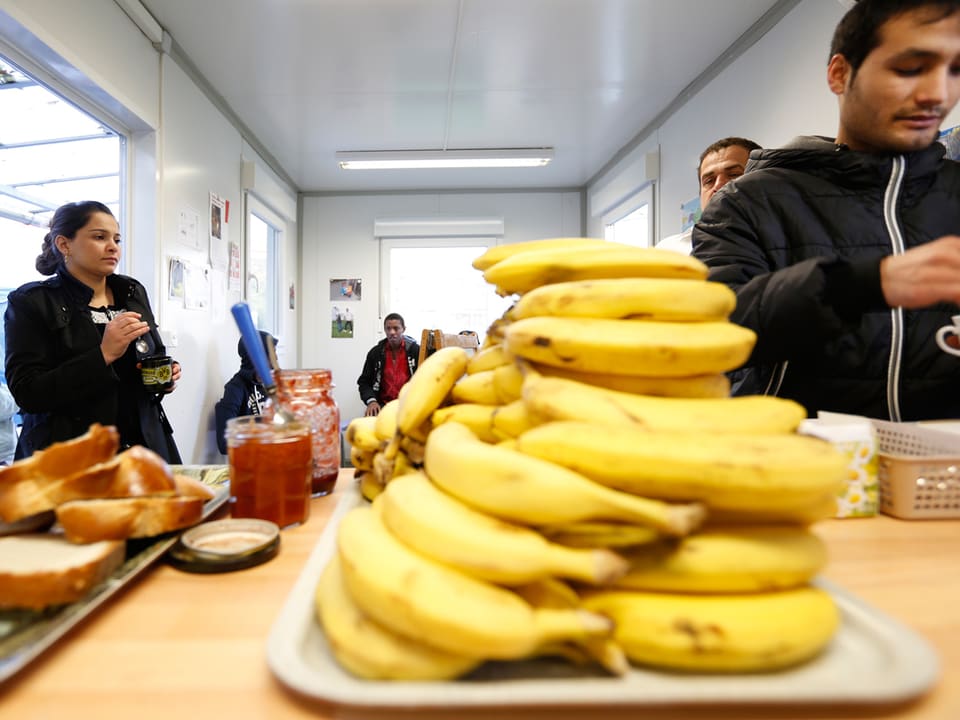 Bananen, Zopfbrot uind Confiture stehen auf einem Tisch, dahinter Menschen.